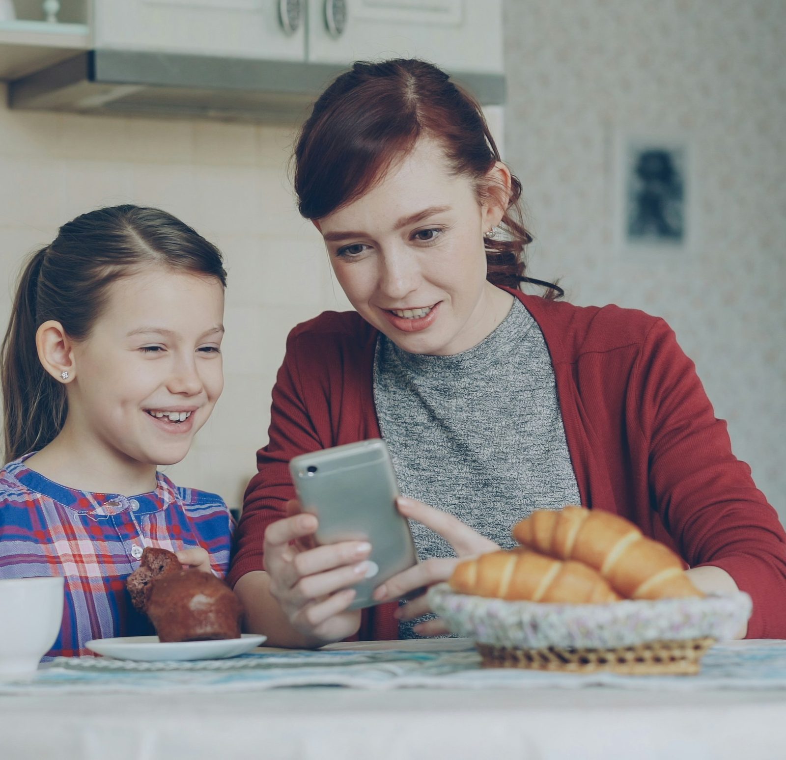 A mother and her daughter smiling at a phone together at breakfast.